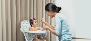 A baby girl in a high chair trying a first solid meal with a spoon fed by her mother